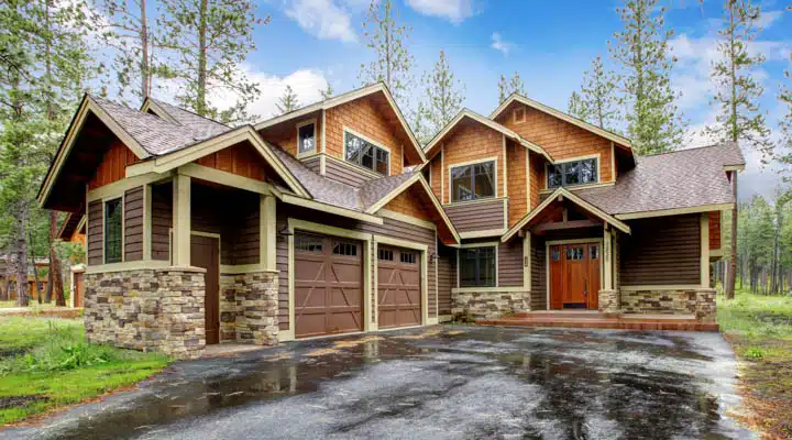 Large mountain cabin house with stone and wet driveway. A large brick building with grass in front of a house
